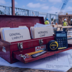 An open red metal toolbox sitting on a blueprint, containing organized file folders labeled 'General Liability' and 'Workers' Comp,' positioned with a modern digital calculator on an active construction site in daylight.
